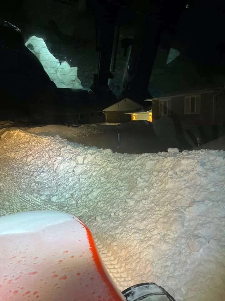 Tractor-cab view of a snowy residential driveway lit by work lights