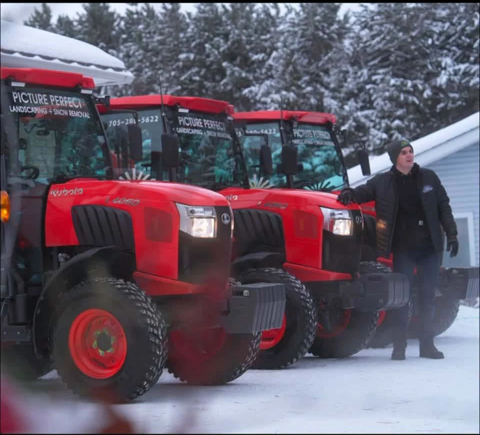 Three orange Picture Perfect tractors lined up in the snow beside a crew member