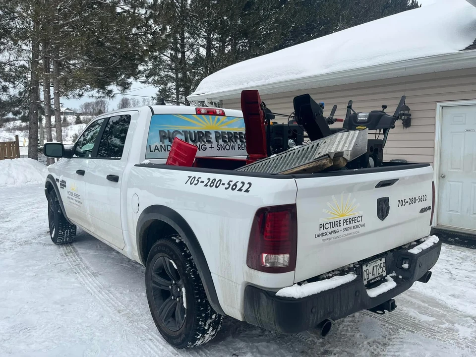 White Picture Perfect pickup truck carrying snow-clearing equipment beside a snowy garage